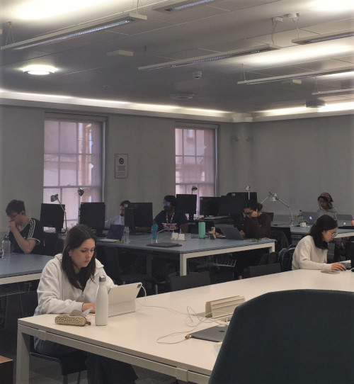 Two students working at a large white desk on their laptops, behind them are other desks with more students, they are working at computers.